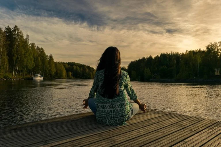 Woman sitting in meditative pose on a wooden dock overlooking a river.