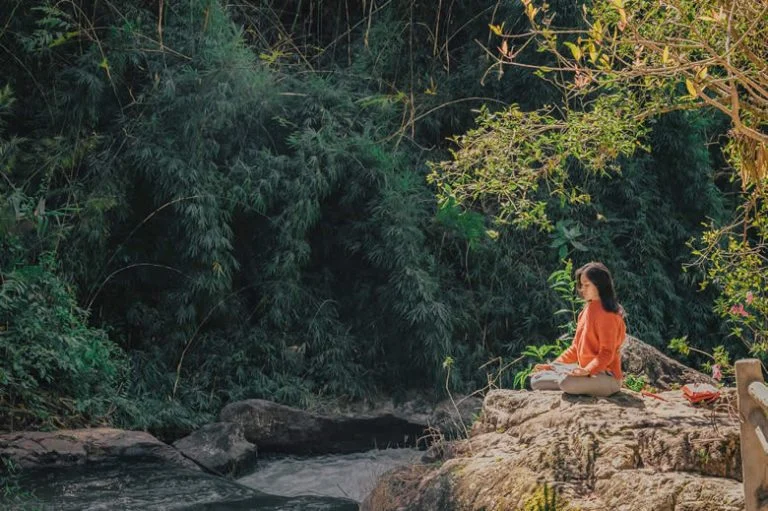 Woman sitting on a rock by a creek meditating.